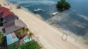 Ein Strand mit klarem Wasser, Booten und einer kleinen Insel. Es sind Gebäude mit roten Dächern und ein Schwimmbad zu sehen.