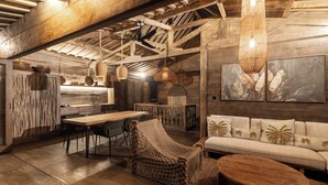 Ein rustikales Interieur mit freiliegenden Holzbalken, einem Essbereich mit Holztisch und -stühlen, einem gewebten Ohrensessel, einer Couch mit dekorativen Kissen und einer hängenden Webdecke.