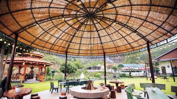 An outdoor patio with a circular fire pit, wooden tables and chairs, and a thatched roof structure.