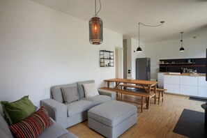 A modern living room with a grey sofa, ottoman, and wooden table. A kitchen with white cabinets and a black countertop is visible in the background. - Up & Up - MyHomeinComo (Moltrasio)
