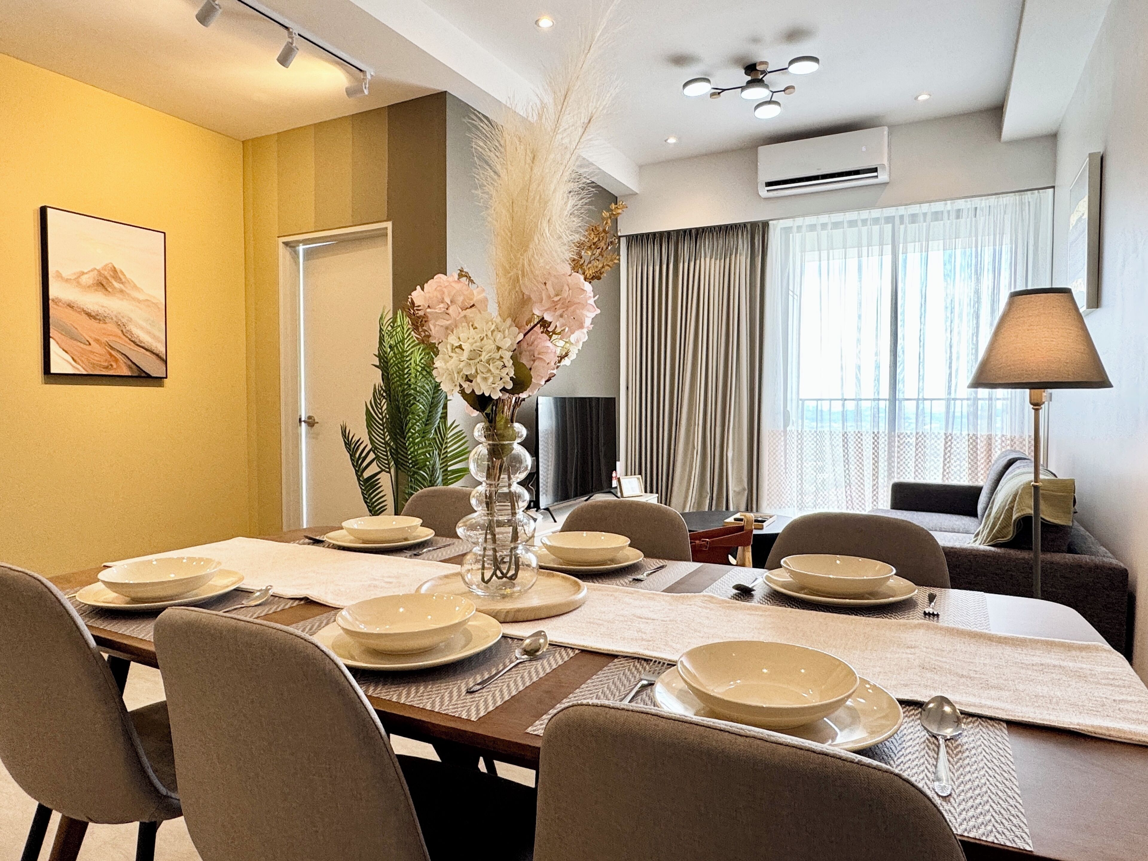 Un comedor con una mesa larga dispuesta para seis personas, rodeada de sillas. Hay un jarrón con flores sobre la mesa, y una ventana grande con cortinas al fondo.
