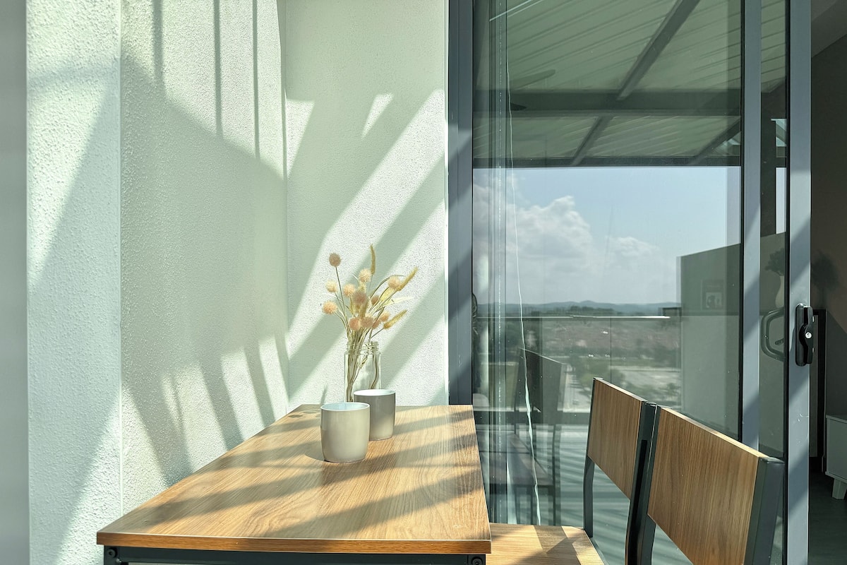 A balcony with a wooden table, a vase with dried flowers, and a glass door leading to a view of the cityscape.