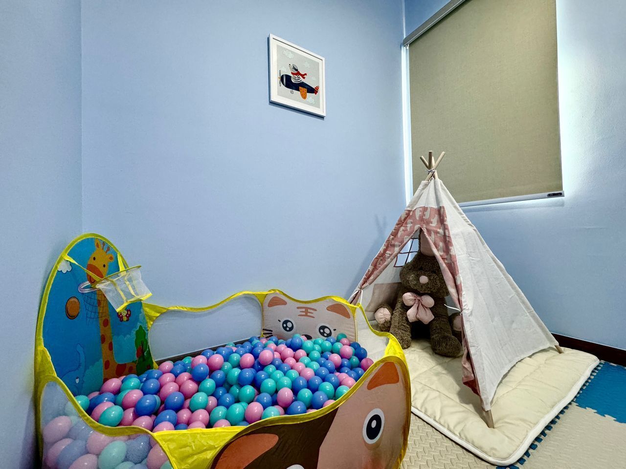 Zona de juegos para niños con piscina de pelotas, alfombra con temática de jirafa, un osito de peluche y un tipi.