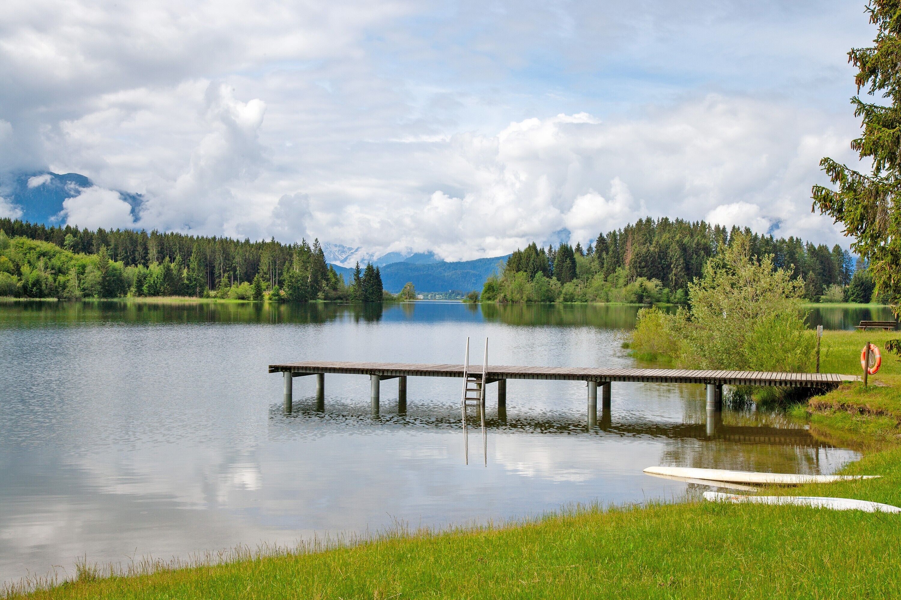 A calm lake with a wooden dock extending into the water, surrounded by lush greenery and distant mountains.