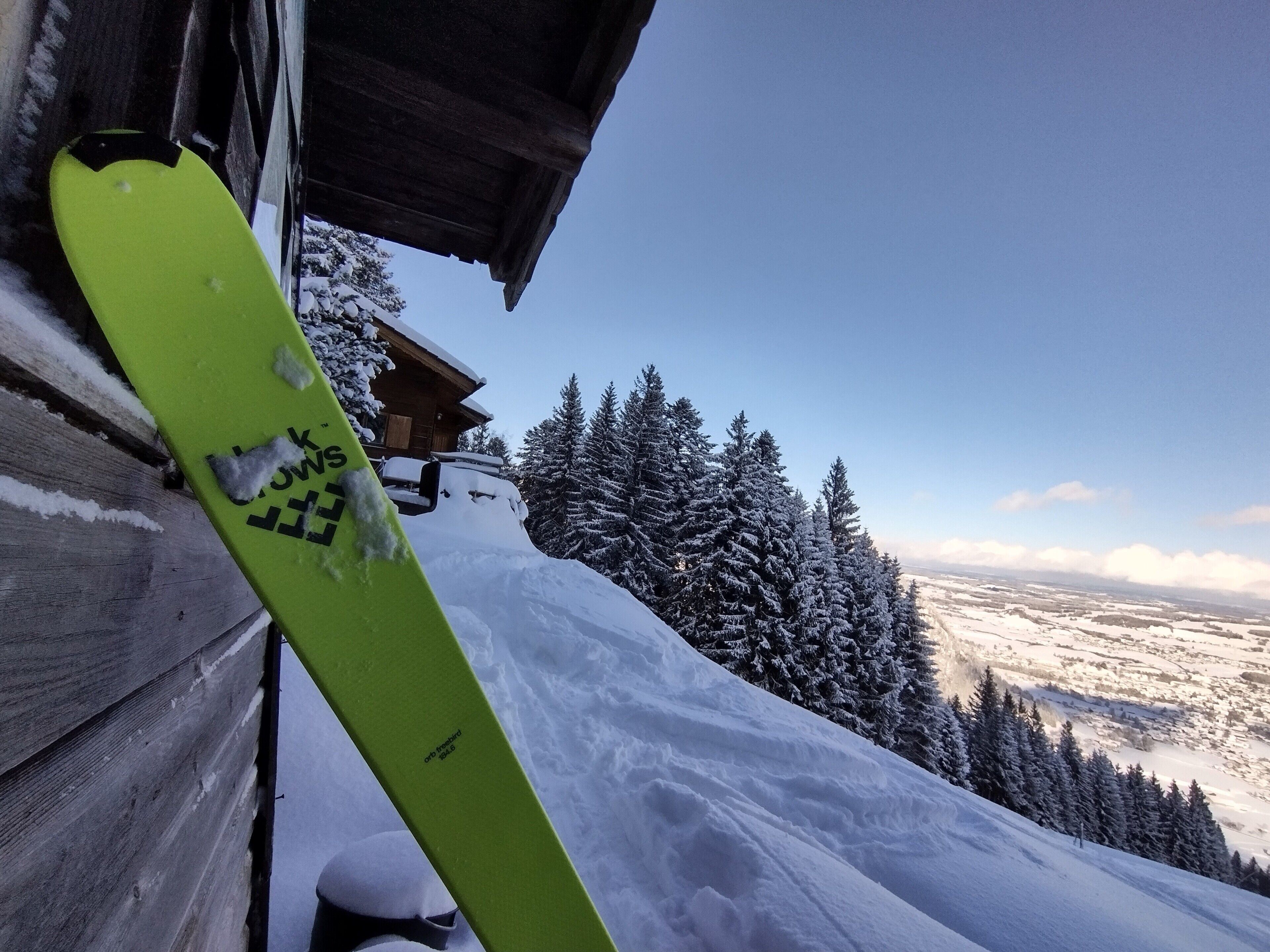 A wooden cabin with a snow-covered roof, a green snowboard leaning against the wall, and a snowy mountain landscape in the background.