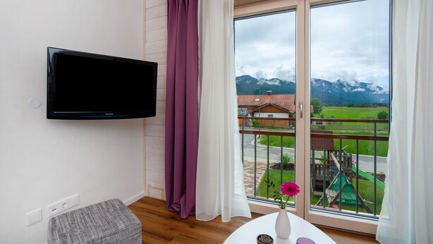 Una habitación con televisión, un taburete pequeño, una mesa con un florero y una taza, y un balcón con vistas a montañas y un parque infantil.