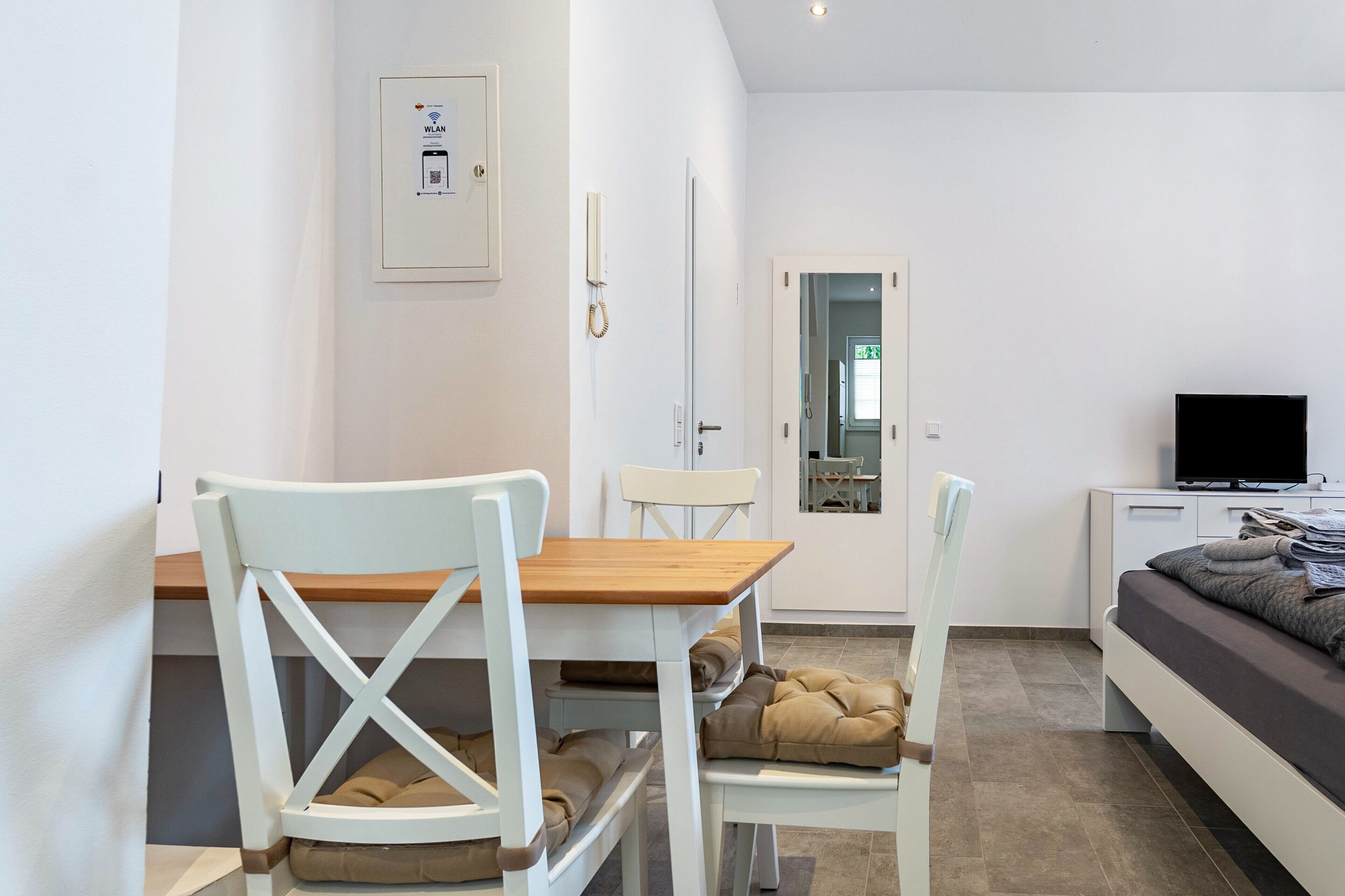 A modern hotel room with a wooden dining table, white chairs, a bed with a grey mattress, a TV on a bedside table, and a door leading to a bathroom.