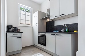 Una cocina moderna con armarios blancos, horno empotrado, estufa, ventana con persianas y una encimera con cafetera y otros electrodomésticos.