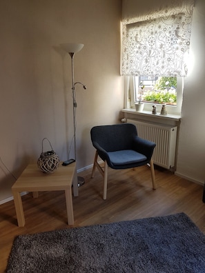 Une pièce avec une chaise bleue, une table en bois, un lampadaire, un lambrequin à motifs et une vue sur la verdure à l’extérieur.