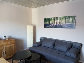 Un salon avec un canapé gris, une table ronde, un meuble et un grand tableau représentant une forêt.
