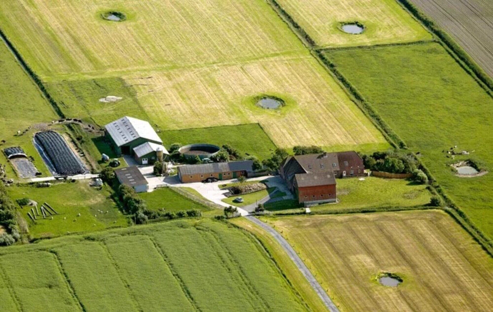 Vista aérea de uma quinta com vários edifícios, incluindo um celeiro grande, estruturas menores e um lago circular.