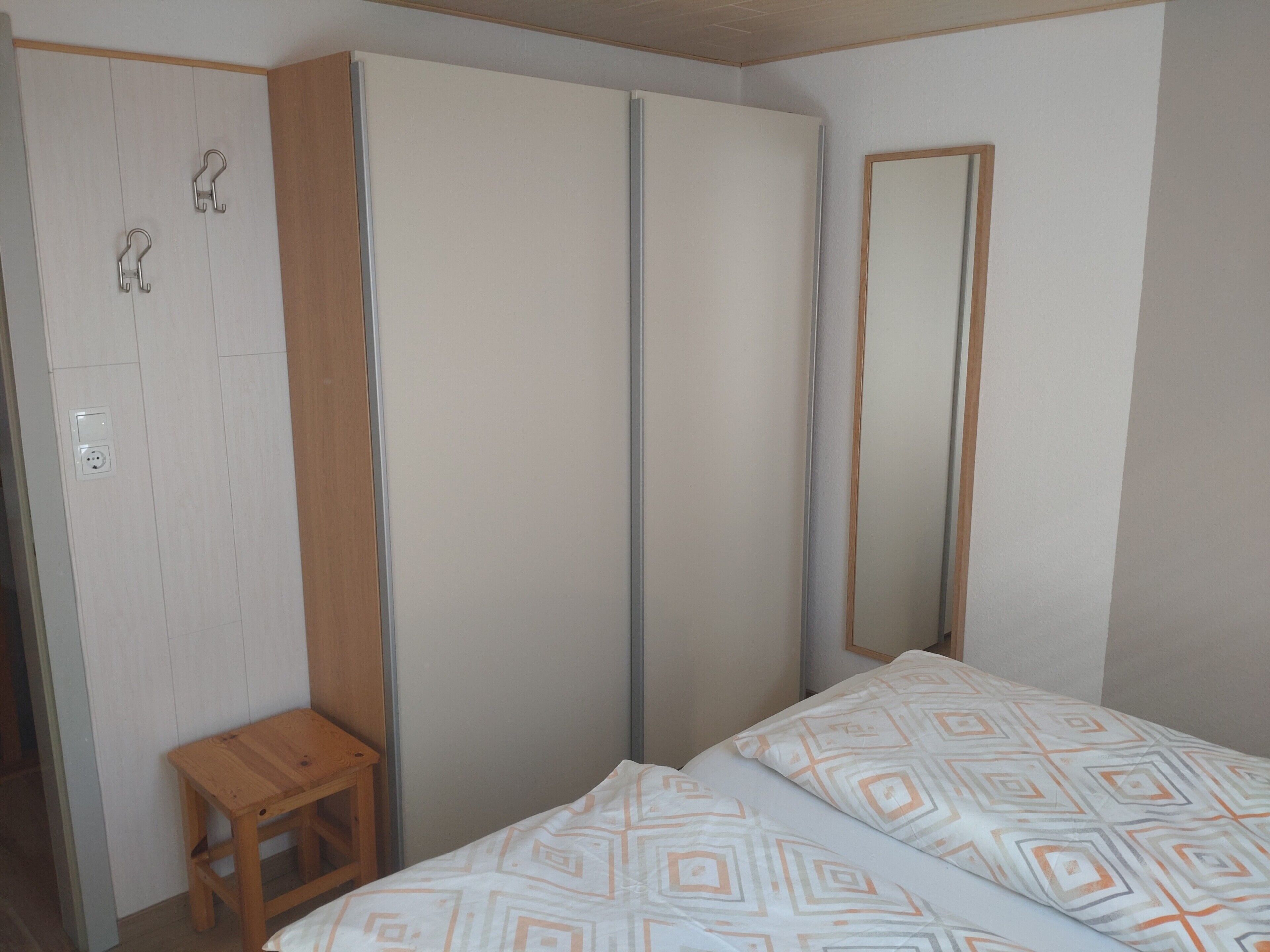 Ein Schlafzimmer mit einem Bett, einem hölzernen Hocker, einem Kleiderschrank und einem Spiegel.