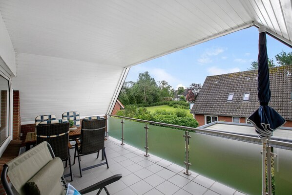 A covered balcony with wicker furniture, a view of a river, and a house in the background. - Apartment 'Große Ferienwohnung' with Shared Garden and Balcony (Nordstrand)