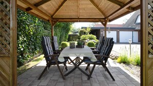 Eine überdachte Terrasse im Freien mit Tisch und Stühlen, umgeben von Pflanzen und einem Gebäude im Hintergrund.