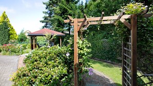 Eine hölzerne Pergola in einem Garten mit üppigem Grün und einem Gazebo im Hintergrund.