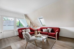 A living room with a red sofa, a glass coffee table, and a white armchair. There is a window with curtains, a radiator, and a skylight.