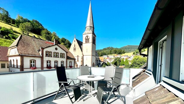 A balcony with a table and chairs, overlooking a town with a church and houses. - Apartment 'Ferienwohnung Schwarzwaldliebe' with Private Terrace and Wi-Fi (Bad Peterstal-Griesbach)
