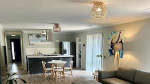 A modern living room with a sectional sofa, a dining area with a small table and chairs, a kitchen with a refrigerator and a sink, and a large window with blinds. - Seaside escape awaits your family in Myalup! (Myalup)