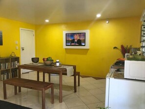 A room with a yellow wall, a television mounted on the wall, a wooden table with chairs, a bench, a bookshelf, and a microwave.