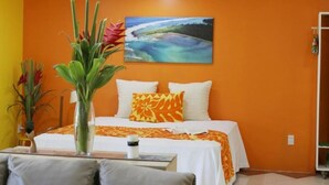 A hotel room with an orange wall, a bed with white and orange bedding, a painting of a coastal scene, and a plant in a vase. - ShineAwayHomes- Mountain view (Matavera District)