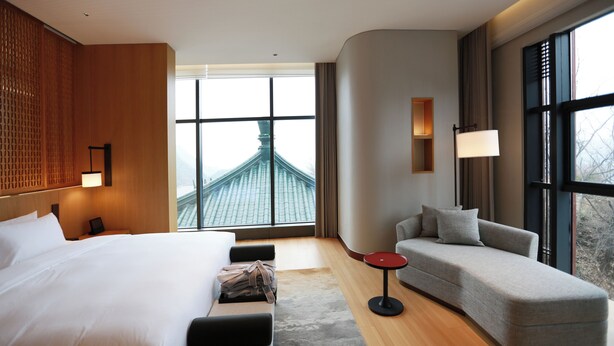 A hotel room with a large bed, a sofa, a small table, and a view of a building through large windows.