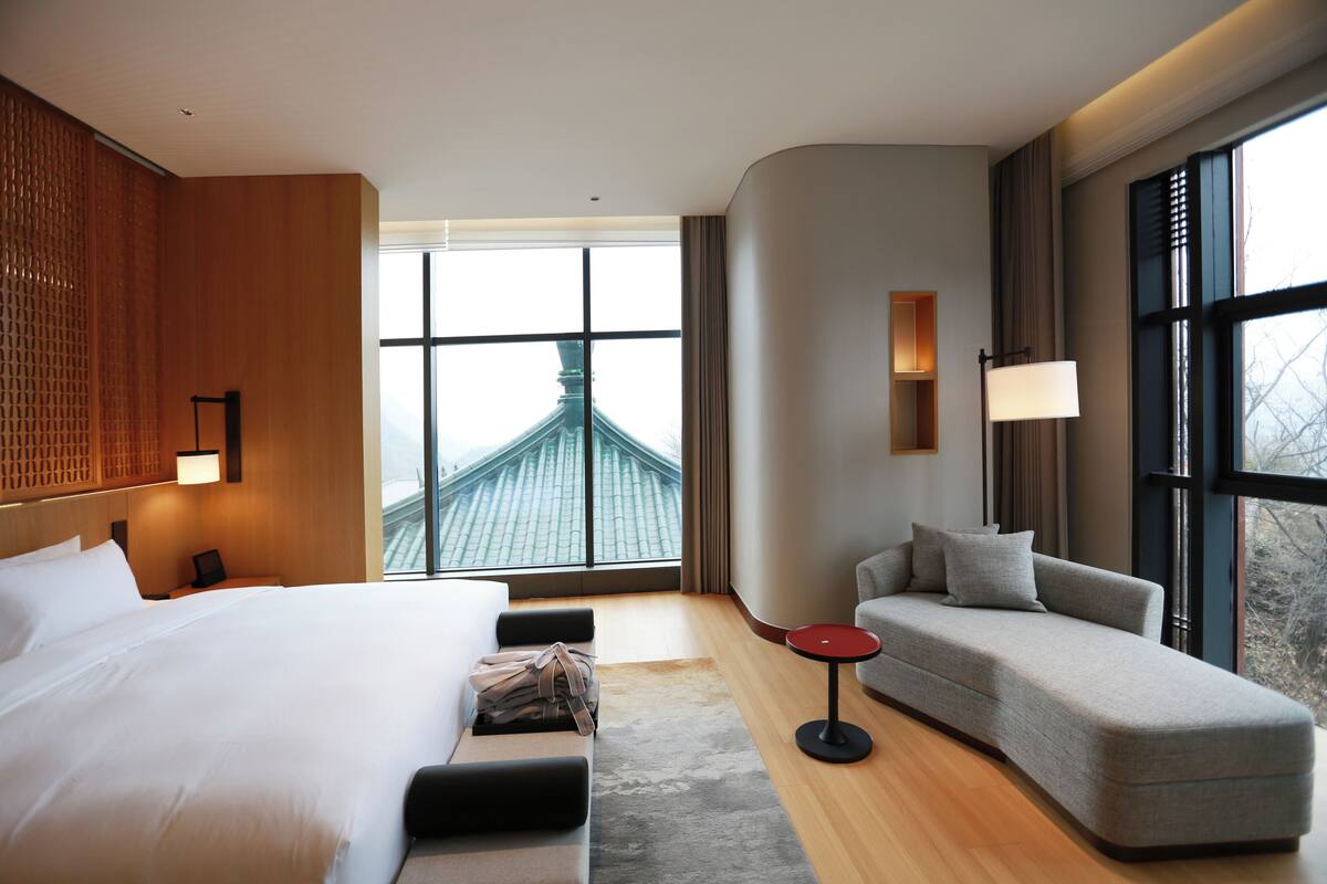 Habitación de hotel con una cama grande, un sofá, una mesita y vistas a un edificio a través de amplias ventanas.