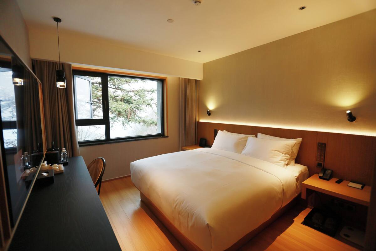 Una habitación de hotel moderna con una cama grande, un escritorio con silla, una ventana con cortinas y una mesita de noche con teléfono y lámpara.