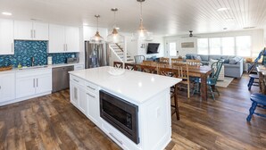 Une cuisine spacieuse avec des meubles blancs, un grand îlot central doté d’un four et un coin repas avec des tables et des chaises en bois.