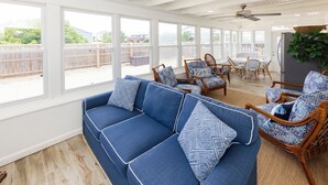 Une véranda spacieuse avec une grande fenêtre, un canapé bleu orné de coussins à motifs, des chaises en osier et une table à manger.