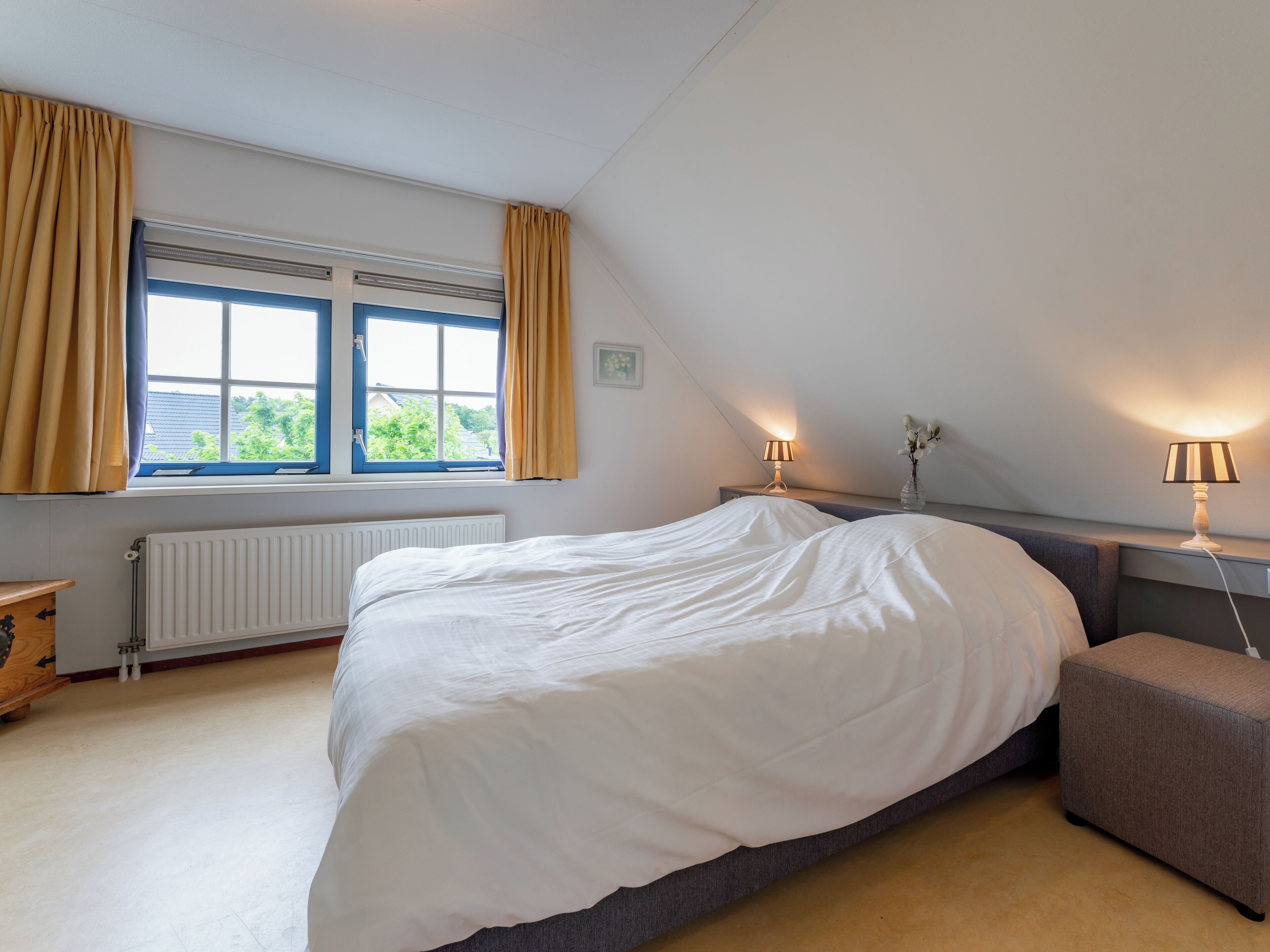 A bedroom with a large bed, a window with curtains, a radiator, a bedside table, and a nightstand with lamps.