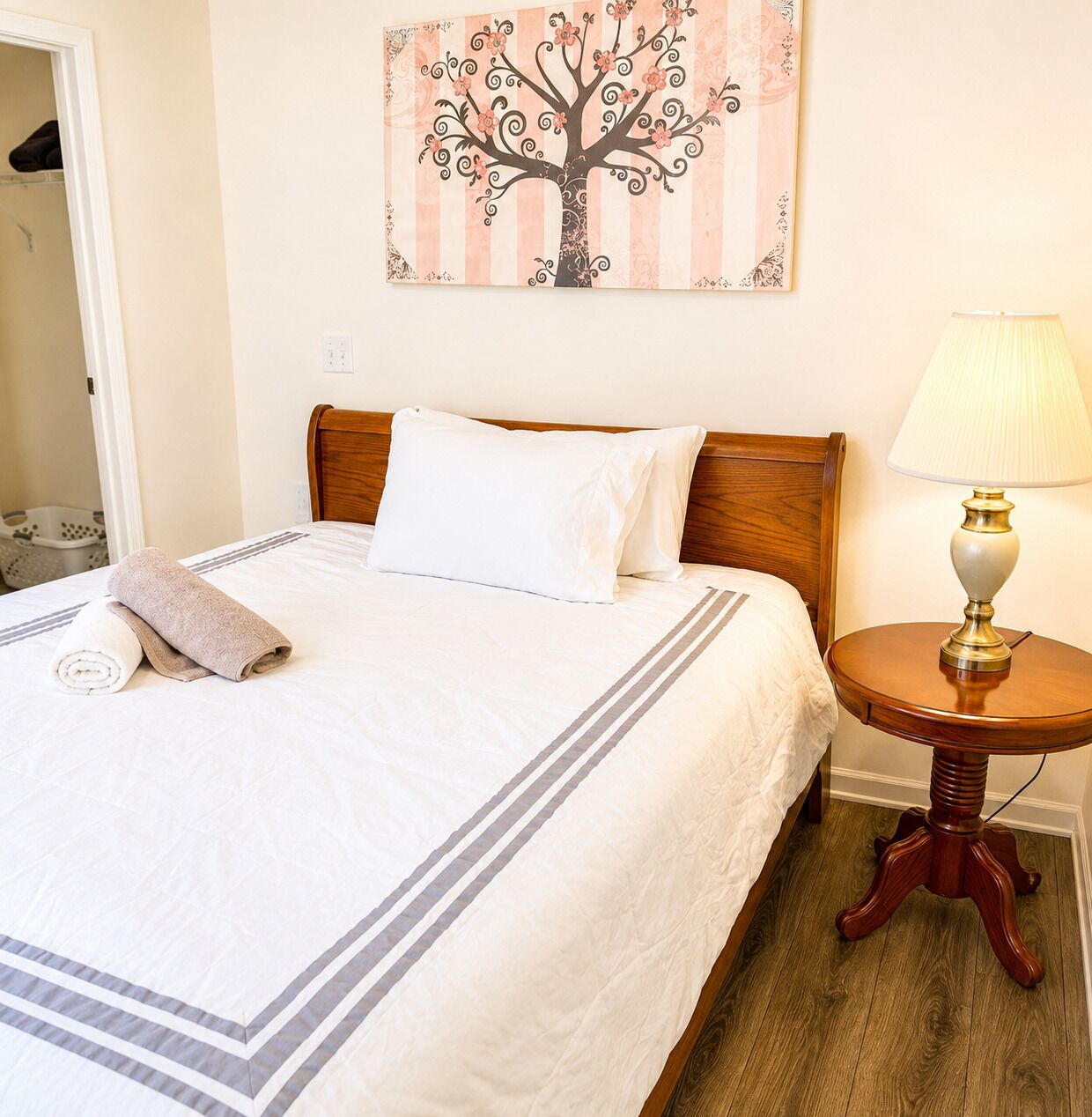 Una cama bien hecha con sábanas blancas y una toalla enrollada, un cabecero de madera, una mesita de noche con una lámpara y un cuadro de un árbol en la pared.