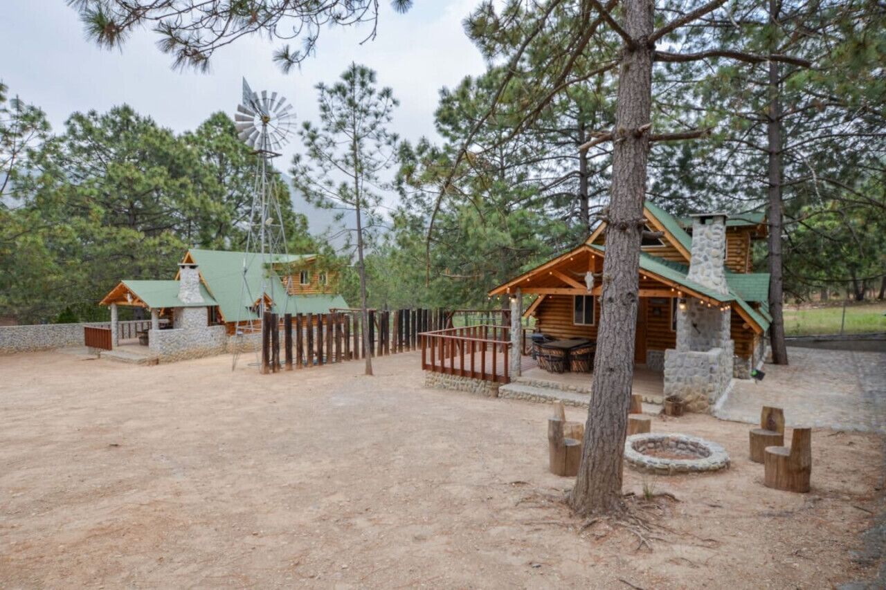 A rustic cabin with a stone chimney, wooden deck, and a fenced area with a fire pit.