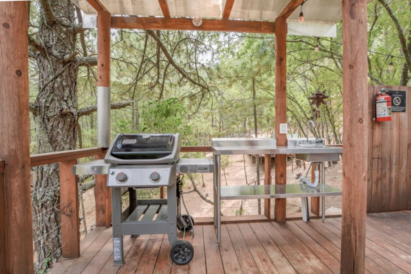 Un área de parrilla al aire libre con una mesa y un extintor sobre una terraza de madera rodeada de árboles.