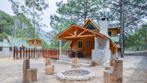 A rustic cabin with a stone chimney, wooden deck, and a fire pit.