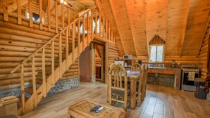 A log cabin interior with a staircase, dining area, kitchen, and a stone fireplace.