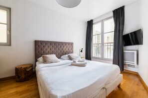 A bedroom with a large bed, a flat-screen TV, a window with curtains, and a wooden floor.