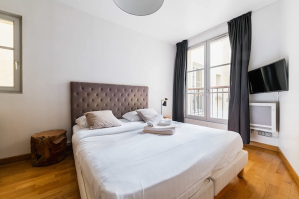 A bedroom with a large bed, a flat-screen TV, a window with curtains, and a wooden floor.