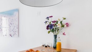 Un comedor con una mesa redonda de madera, dos sillas negras, una lámpara colgante gris, un jarrón con flores, una botella de jugo de naranja y un plato de cruasanes.