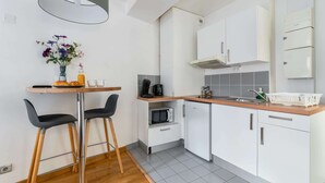 Una cocina compacta con gabinetes blancos, microondas, cafetera y un refrigerador pequeño. Hay una mesa de madera con dos sillas y un jarrón con flores.