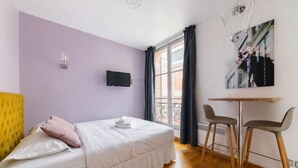 A bedroom with a bed, a desk, two chairs, a TV, and a window with curtains.