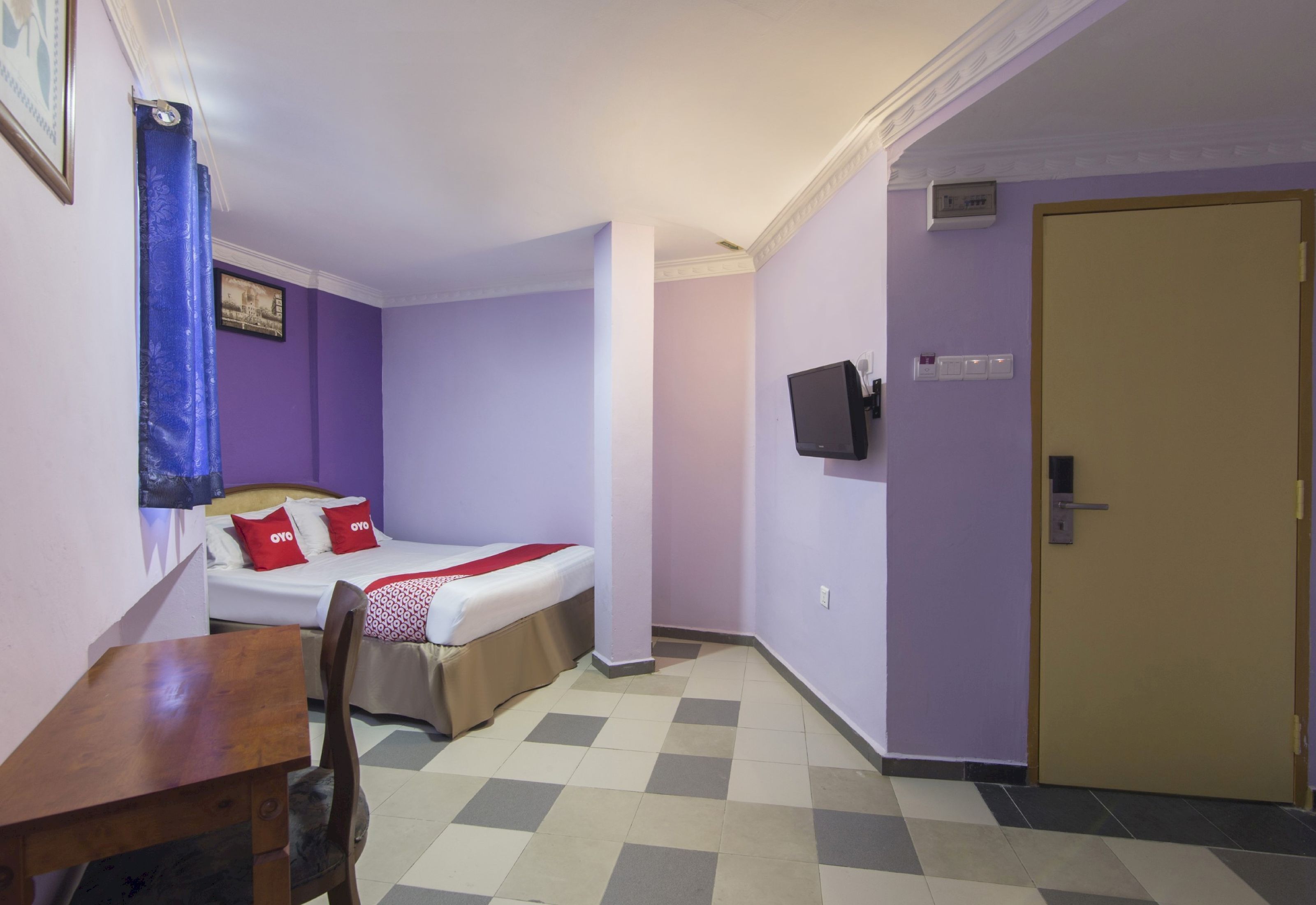 Una habitación pequeña y ordenada con una cama, un escritorio y una silla. Se observa una puerta y un televisor montado en la pared.