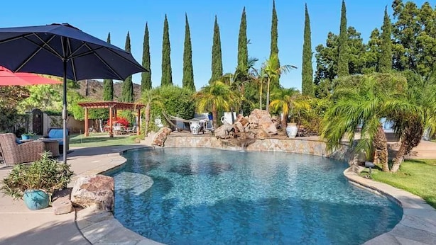 Una piscina con borde de piedra, rodeada de palmeras y cipreses.