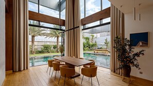 A modern dining area with a wooden table and chairs, large windows, and a view of a swimming pool and greenery. - Glamorous getaway VILLA in Bien Hoa (Bien Hoa)