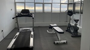 Ett gym med löpband, crosstrainer och en träningsbänk. Det har stora fönster med utsikt över himlen och molnen.