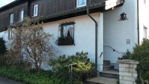 A two-story building with a dark wooden facade and white walls. There are two windows on the upper floor and a small balcony with a metal railing. - Ferienwohnung Lehmann 58 qm, Erdgeschoss, mit Terrasse und Separatem Schlafzimmer (Bad Endorf)