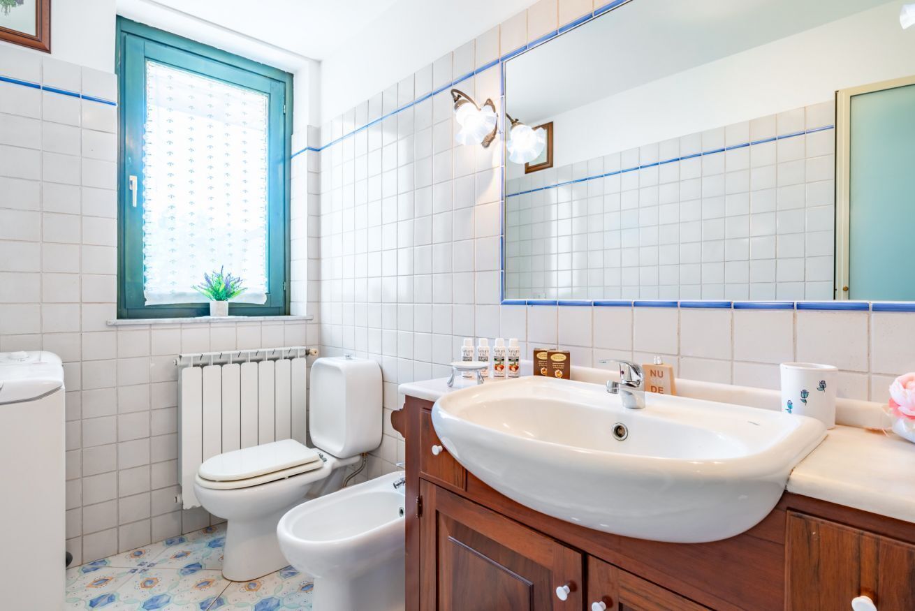 Une salle de bain comprenant des toilettes, un bidet, un lavabo, un miroir et une fenêtre avec des stores.