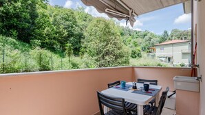 En balkon med et bord og stole, en lille vask og udsigt til en grøn bakke med træer og bygninger.