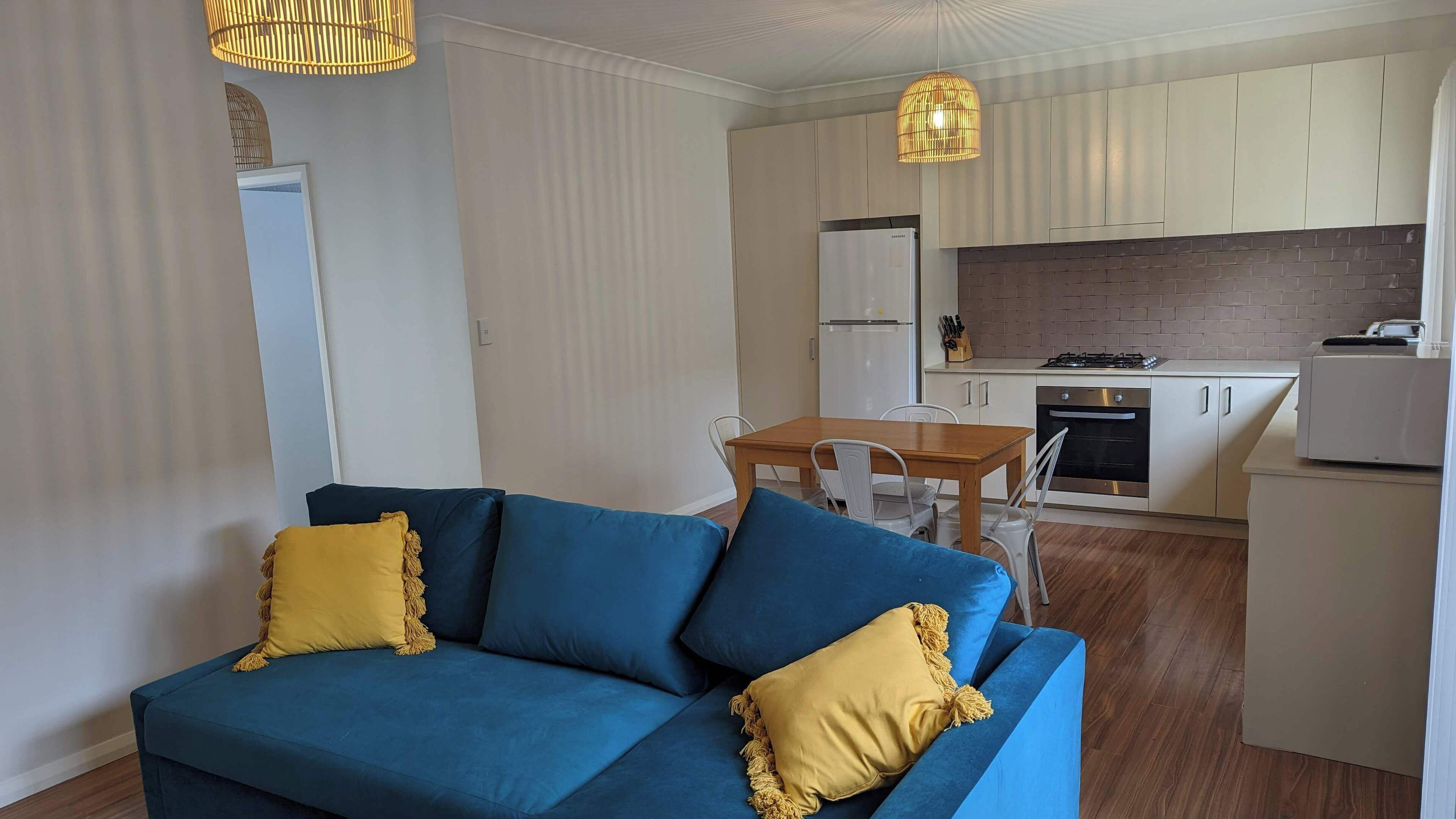 A modern living room with a blue sofa and yellow cushions, a wooden dining table, and a kitchen with white cabinets and a brick backsplash.