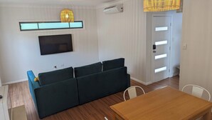 A modern living room with a sofa, a TV, a wooden dining table, and a door with a light switch. - 1 bedroom guesthouse near airport with private access and parking (Arncliffe)