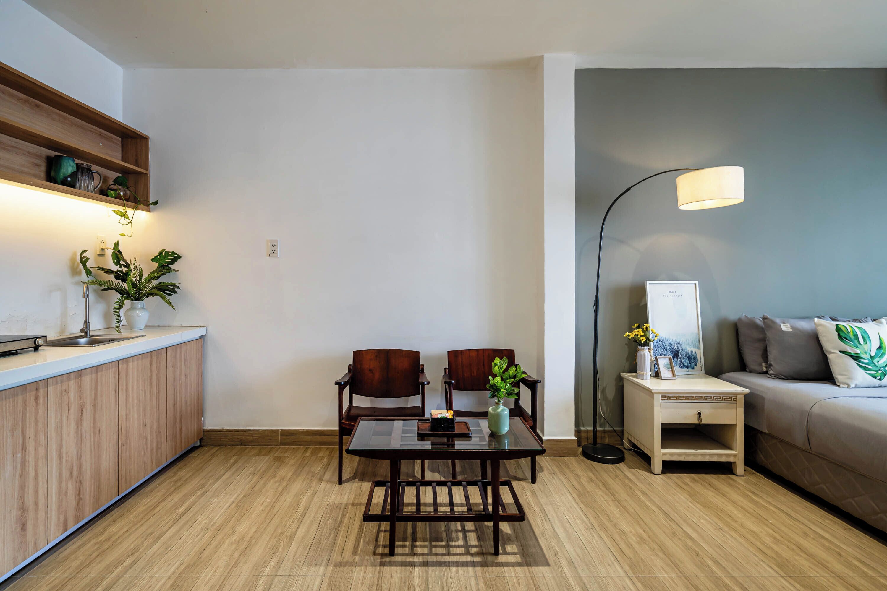 Un salon moderne avec un plancher en bois, une table à manger avec un plateau en verre, deux chaises en bois, un canapé, une table d’appoint avec une lampe et un petit meuble.